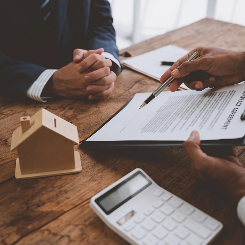 Signature d'un contrat immobilier 'CONTRACT AGREEMENT' avec stylo. Un modèle de maison en bois et une calculatrice sont sur la table en bois.